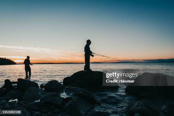 silhouettes of a mother with son, fishing from large stones with spinning rods in the sea at twilight - fisherman isolated stock pictures, royalty-free photos & images