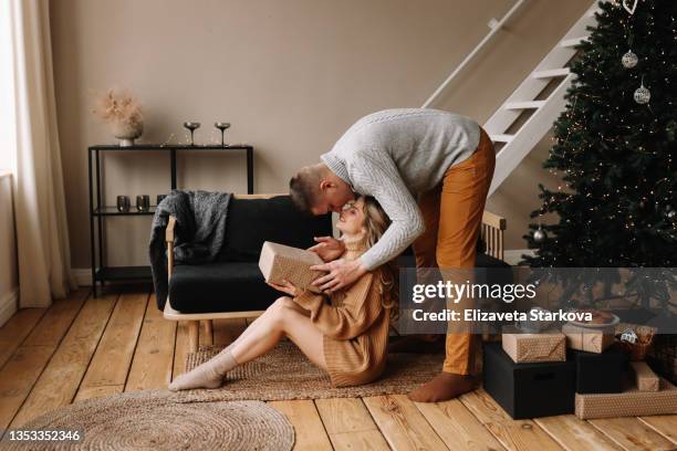 a man gives a christmas gift box to a woman in a decorated cozy room with a christmas tree at home on the new year's holiday indoor. a couple in love celebrates christmas together - christmas couple stockfoto's en -beelden