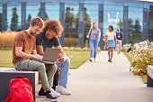 Male University Or College Students Sitting Outdoors On Campus Talking And Working On Laptop