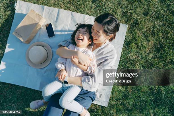 affectionate young asian mother embracing little daughter in arms, lying down on the grassy field, having fun and smiling joyfully, enjoying together on a sunny day. family love and bonding time. enjoying the nature. outdoor fun concept - asia fotografías e imágenes de stock