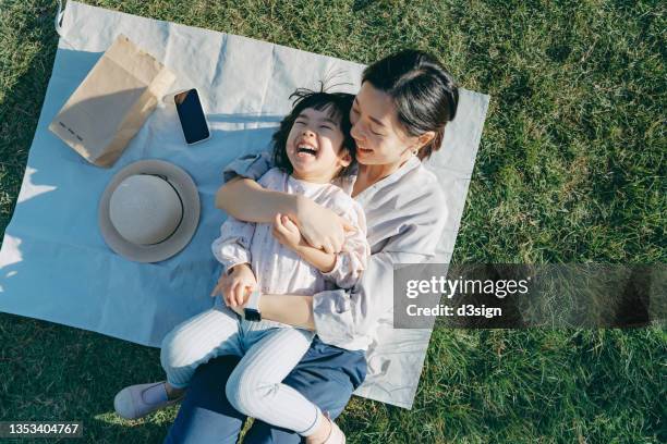 affectionate young asian mother embracing little daughter in arms, lying down on the grassy field, having fun and smiling joyfully, enjoying together on a sunny day. family love and bonding time. enjoying the nature. outdoor fun concept - asian photos et images de collection