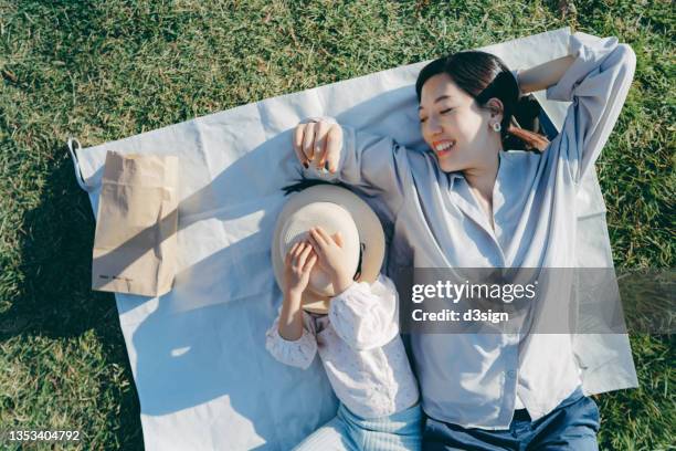 affectionate young asian mother and little daughter lying down on the grassy field, having fun playing and smiling joyfully, enjoying together on a sunny day. family love and bonding time. enjoying the nature. outdoor fun concept - family no faces stockfoto's en -beelden