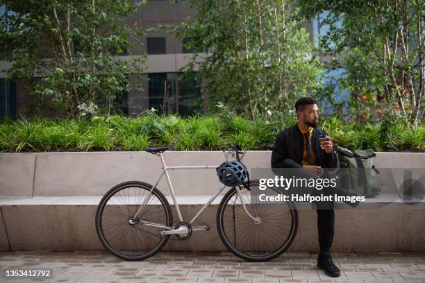 young man commuter with bicycle sitting and drinking coffee on bench outdoors in city. - bomberjacka bildbanksfoton och bilder