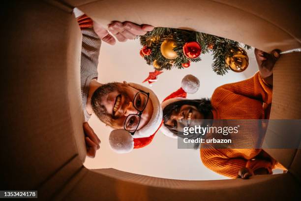 multiethnisches paar packt weihnachtsgeschenk aus - nikolausmütze stock-fotos und bilder