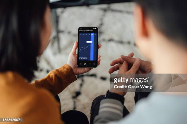 over the shoulder view of young couple managing financial investment with mobile banking app on smart phone - couple smartphone stockfoto's en -beelden