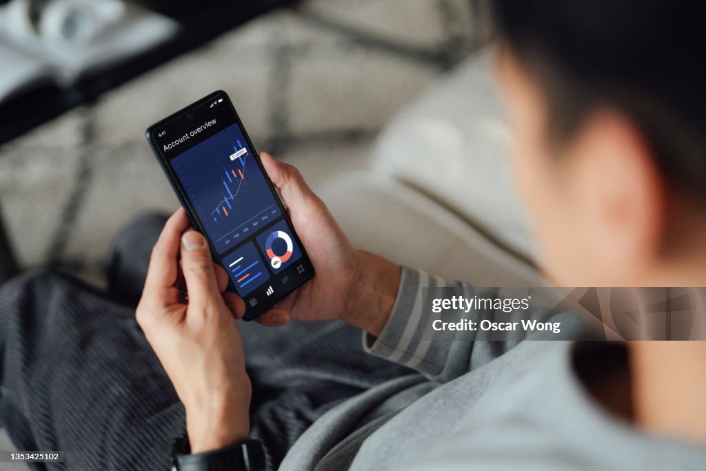 Over the shoulder view of young man checking financial data with mobile banking app on smart phone while sitting on sofa at home