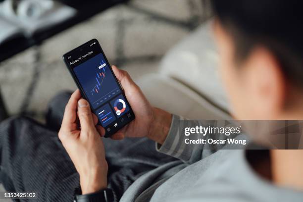 over the shoulder view of young man checking financial data with mobile banking app on smart phone while sitting on sofa at home - look back stock pictures, royalty-free photos & images