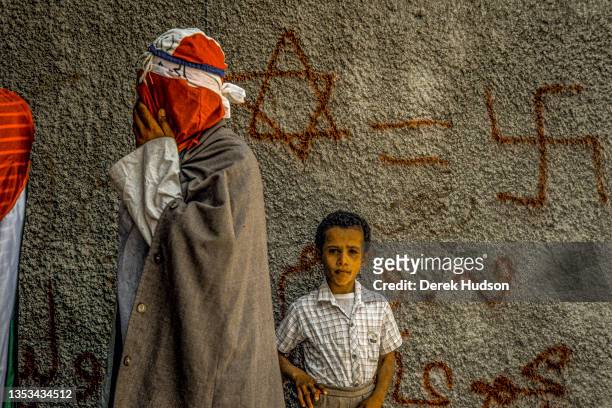 View of an unidentified young boy as he stands in front of a graffiti-covered wall, as a masked, Pro-Palestinian activist walks past, during the...