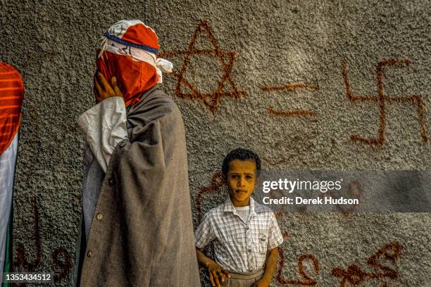 View of an unidentified young boy as he stands in front of a graffiti-covered wall, as a masked, Pro-Palestinian activist walks past, during the...