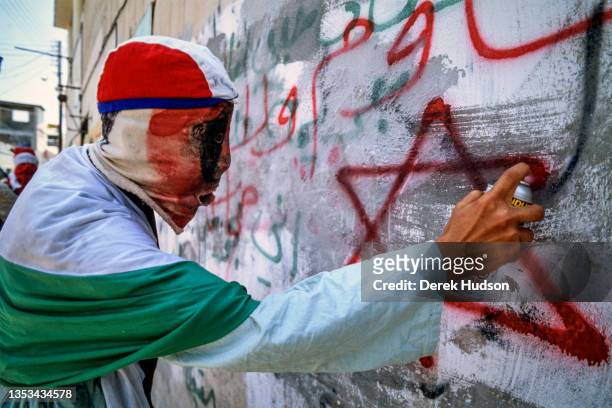 Profile view of an unidentified Pro-Palestinian activist, wrapped in the colors of the Palestinian flag, spray painting to graffiti a wall during the...