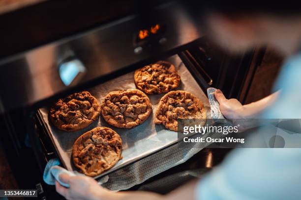 over the shoulder view of young woman taking out freshly baked cookies from the oven - faire cuire au four photos et images de collection