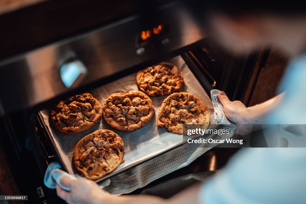 Over the shoulder view of young woman taking out freshly baked cookies from the oven
