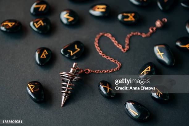 bronze dowsing pendulum and many runes made of obsidian with gold text arranged on black background. concept of magic esoteric rituals. macro photography in flat lay style - ónix fotografías e imágenes de stock