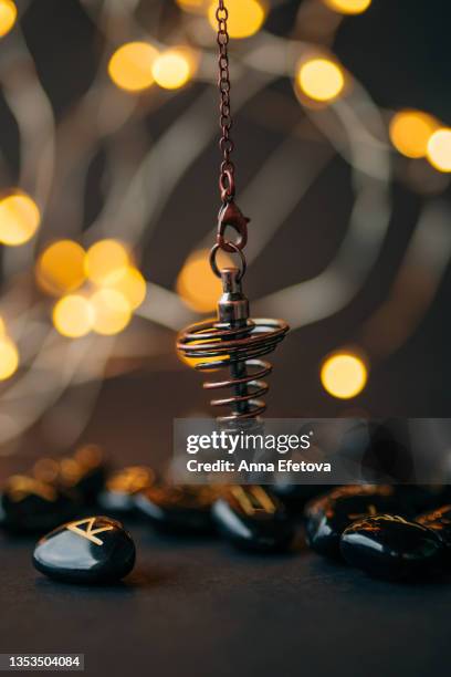 bronze dowsing pendulum and runes made of obsidian with gold text with garland lights background. concept of magic esoteric rituals. copy space for your design - rune symbols stock pictures, royalty-free photos & images