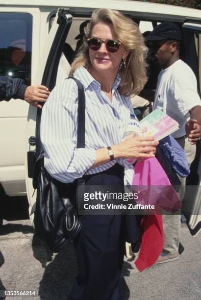 American actress Candice Bergen wearing a blue-and-white striped blouse and sunglasses, carrying a bottle of water and a book, on which is written 'A...