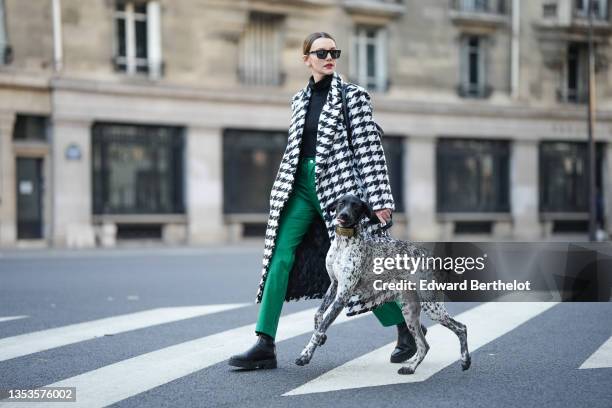 Olesya Senchenko wears black sunglasses, gold earrings, a black wool turtleneck pullover, a black and white large houndstooths print pattern long...