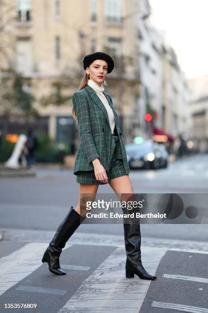 Olesya Senchenko wears a black felt / wool beret, gold earrings, a white ribbed wool turtleneck pullover, a green and white checkered print pattern...