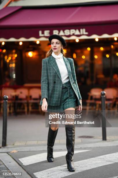Olesya Senchenko wears a black felt / wool beret, gold earrings, a white ribbed wool turtleneck pullover, a green and white checkered print pattern...