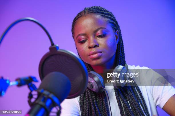 a young dark-skinned girl in a white t-shirt with afro-braids is looking down, sitting in a recording studio with violet background - radio dj stock pictures, royalty-free photos & images