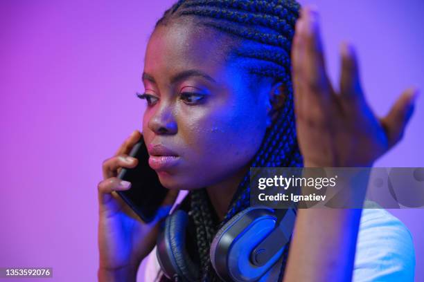 a distraught young african woman with headsets on her neck talks on the phone and gesticulating in a recording studio with violet background - mobile phone edit stockfoto's en -beelden