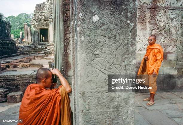 An monk , in a saffron-colored, takes a photograph of a second monk, in a turmeric-colored robe, who poses at the Angkor Wat, temple complex, Siem...