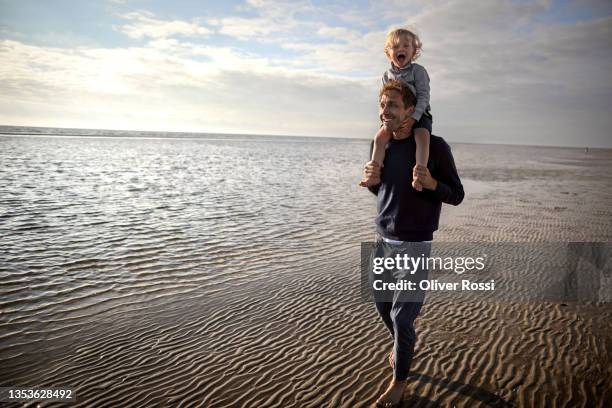 happy father carrying son on shoulders on the beach - llevar al hombro fotografías e imágenes de stock