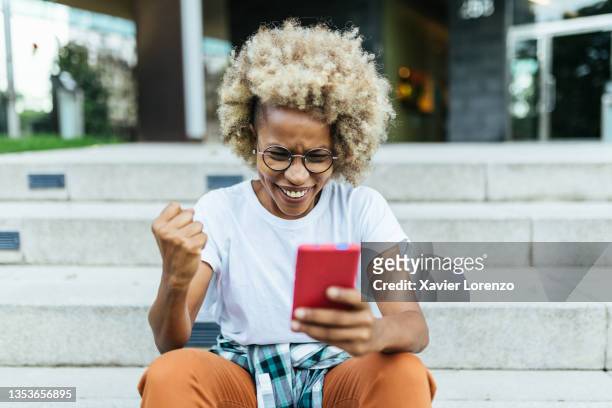 happy african american woman celebrating victory while receiving good news on her mobile phone. - cheering stock-fotos und bilder