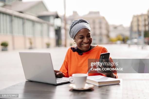 woman using a mobile phone and a computer laptop while sitting at a coffee shop outdoors. - african american restaurant texting stockfoto's en -beelden