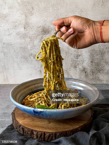 image of metal fork twirling pesto, spaghetti held in hand of unrecognisable person, pasta topped with homemade pesto sauce, portion on blue and white dish garnished with basil leaves, elevated view, focus on foreground - pesto imagens e fotografias de stock