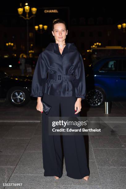 Ilona Smet wears diamond earrings, a black silk puffy sleeves / high collar / belted coat, black high waist palazzo / large pants, a black shiny...