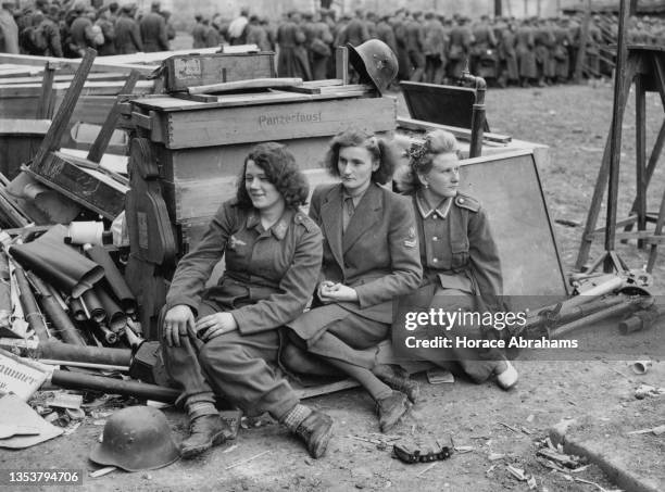 Three female German Luftwaffe and Wehrmachthelferin prisoners of war sit amidst abandoned weapons and ammunition boxes after surrendering to units of...