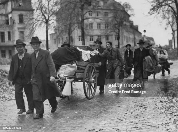 Homeless German civilian refugees carry their bundles of clothes and possessions in suitcases and hand pushed carts past abandoned bomb damaged...