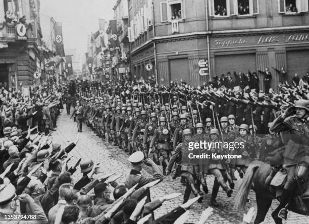Crowds of people give the Nazi salute as German Wehrmacht troops goose step march through the town square of Friedland in the occupied Sudetenland of...