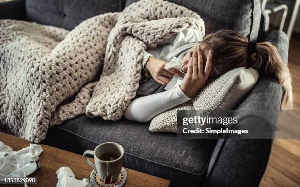 very sick young woman lies on a couch holding her forearm and handkerchief at her nose fighting fever. - blocked nose stock-fotos und bilder