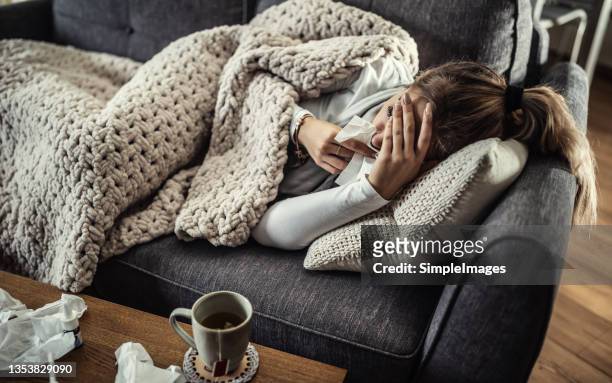 very sick young woman lies on a couch holding her forearm and handkerchief at her nose fighting fever. - état grippal photos et images de collection