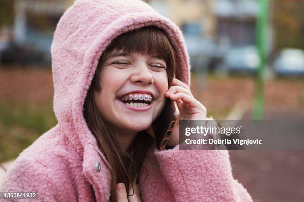 adorable girl with eyes closed and dental braces laughing when having a phone conversation outdoors - chatting park stockfoto's en -beelden