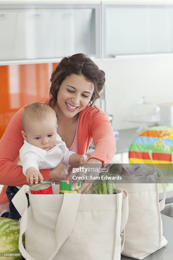 Woman holding baby and unloading groceries from reusable bag