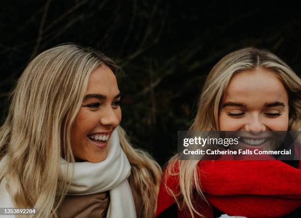 a portrait of two happy, beautiful young blond woman sitting outdoors, wearing scarves and coats. one woman looks over at the other. - autumn friends coats stock pictures, royalty-free photos & images