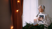 Nostalgic senior woman holding picture frame, standing by window at home.