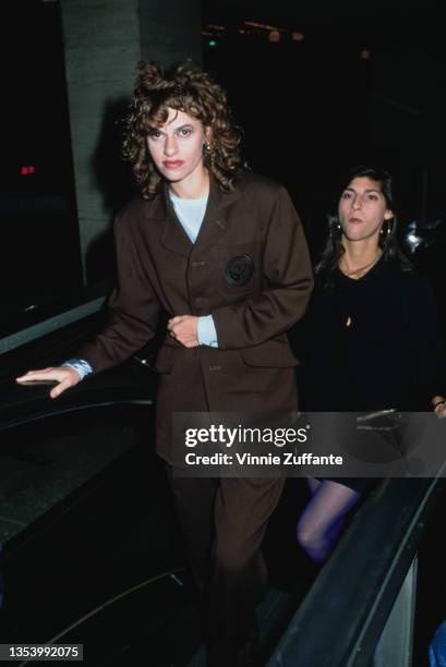 American actress and comedian Sandra Bernhard wearing a dark brown suit, buttoned up over a pale blue top, as she reaches the top of an escalator,...