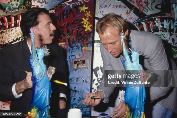 German-born American artist Peter Max and American actor Corbin Bernsen holding paintbrushes at a table on which there are figurines of the Statue of...