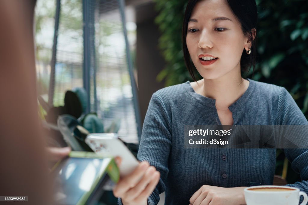 Close up of young Asian woman making mobile payment with her smartphone in a restaurant, scan and pay a bill on a card machine making a quick and easy contactless payment. NFC technology, tap and go concept
