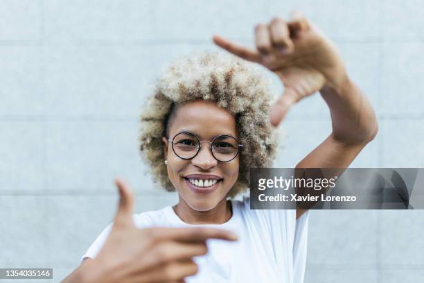 cheerful african american woman looking at camera and smiling while shaping frame with her fingers in front of wall in city street - identity stock-fotos und bilder