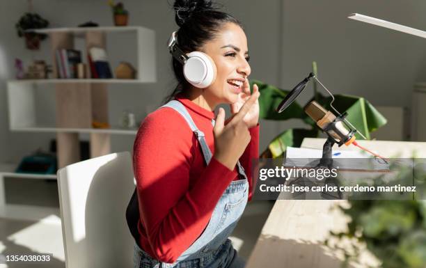 woman sitting at home desk gestures while talking into microphone and wearing headphones recording a podcast - multimedia stockfoto's en -beelden