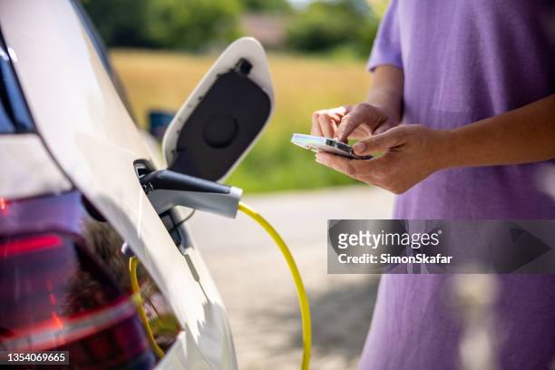 woman using mobile phone while charging electric car - electric car home stock pictures, royalty-free photos & images