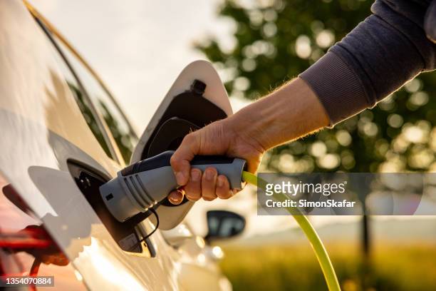 man inserts a power cord into an electric car for charging in the nature - part of stock pictures, royalty-free photos & images