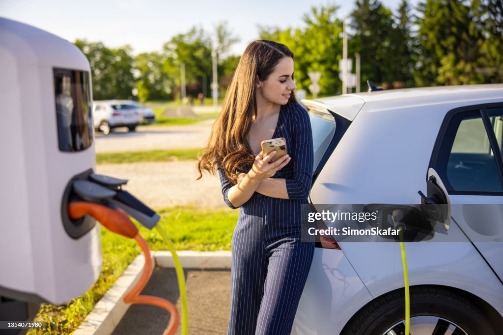 Junge Frau mit Handy in der Hand, während sie auf das Aufladen des Elektroautos wartet
