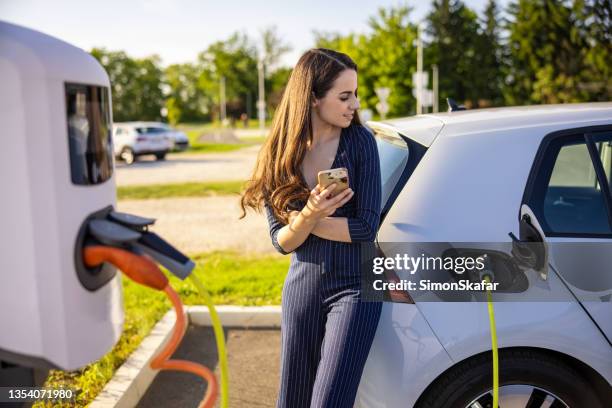 junge frau mit handy in der hand, während sie auf das aufladen des elektroautos wartet - hybrid vehicle stock-fotos und bilder