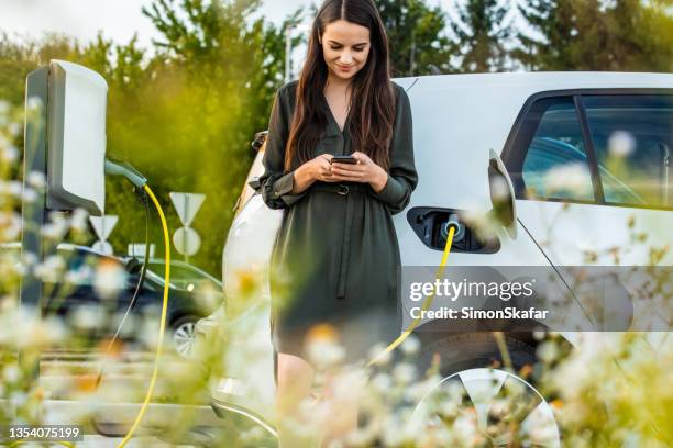 woman using mobile phone while waiting for electric car to charge in the parking lot - electric cars stockfoto's en -beelden