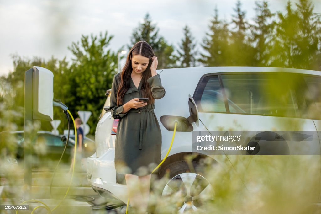 Frau mit Handy, während sie auf Elektroauto auf dem Parkplatz aufladen